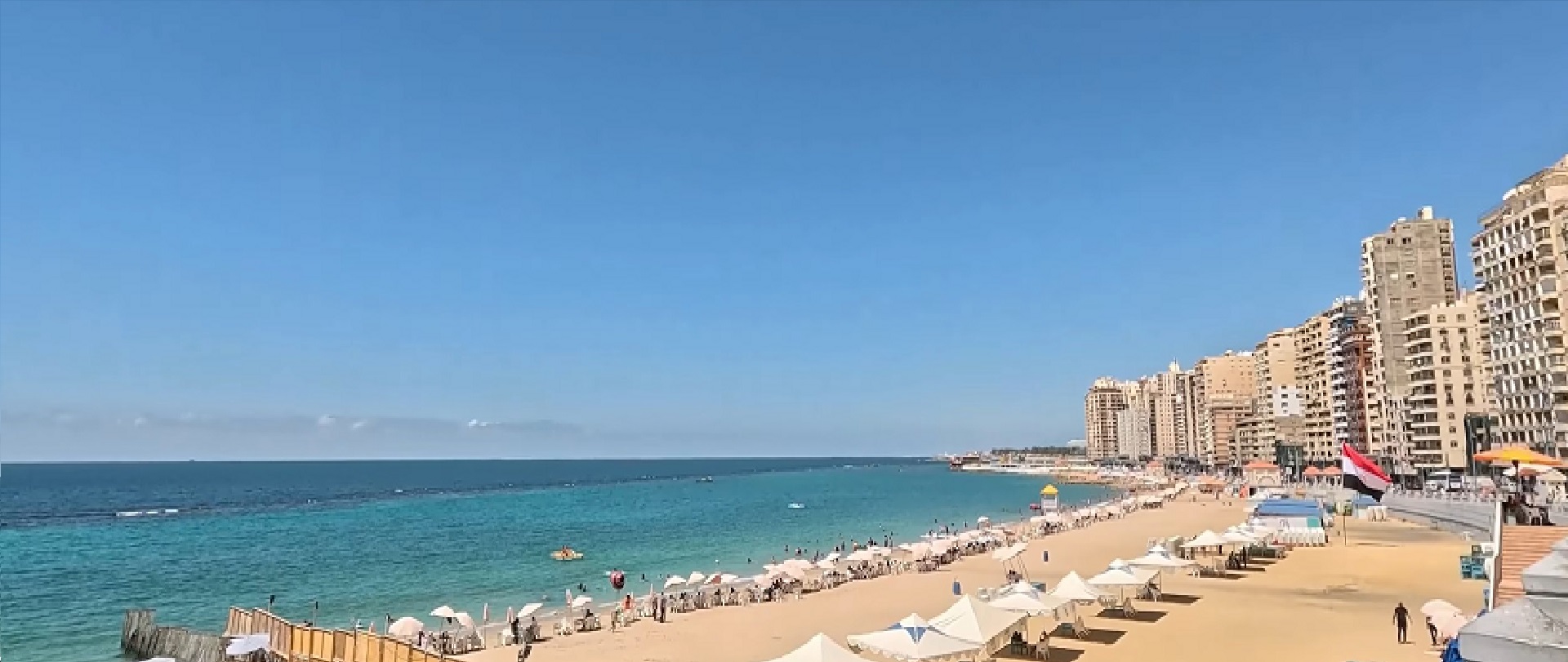 Spiaggia con ombrelloni bianchi e palazzi sulla Corniche di Alessandria, mare turchese