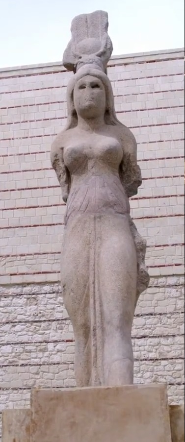 Eine verwitterte Steinstatue einer Frau mit kunstvollem Kopfschmuck vor einer Ziegelwand